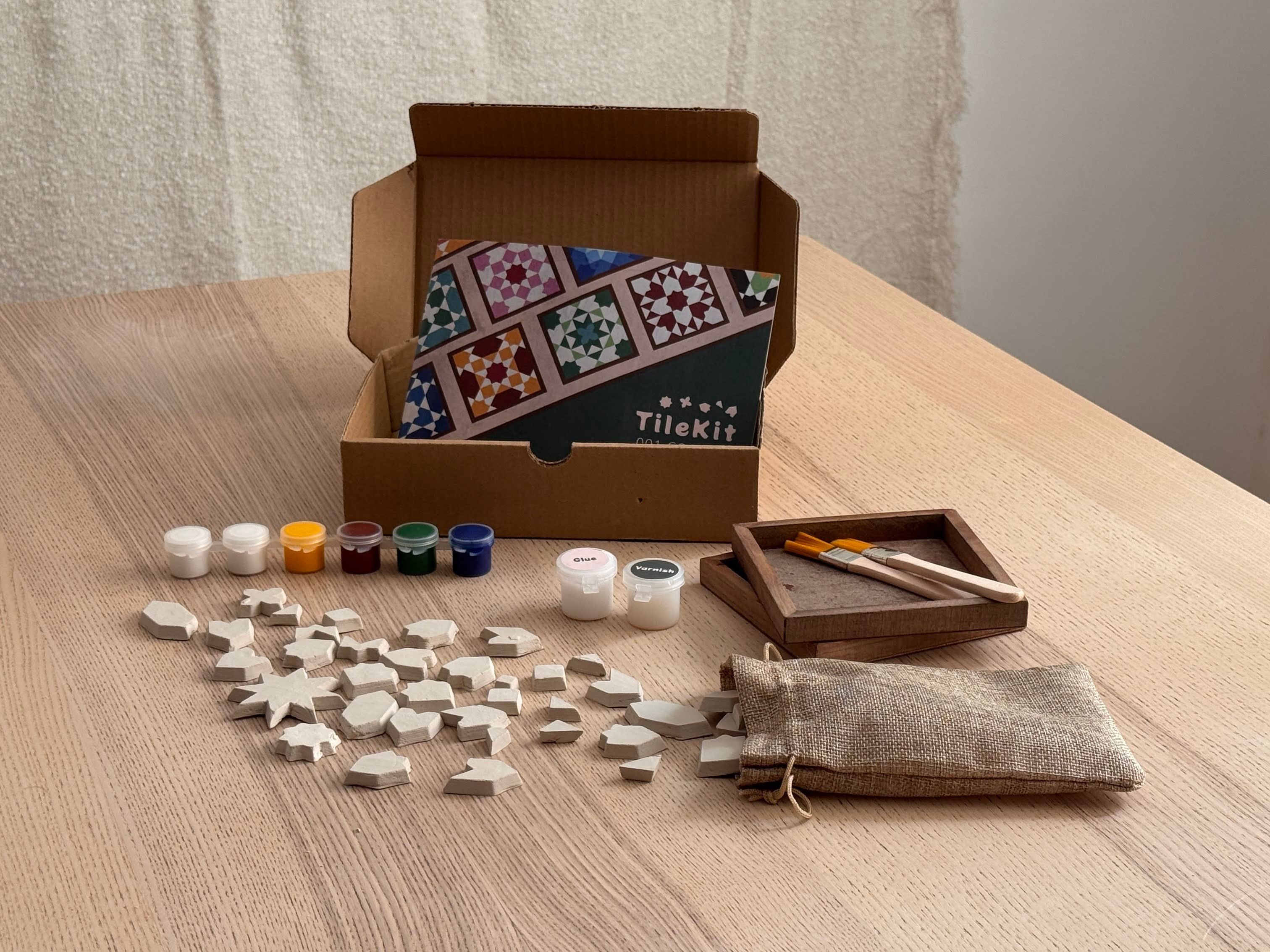 TileKit box open on a table with clay tiles, paints, brushes and coaster bases laid out
