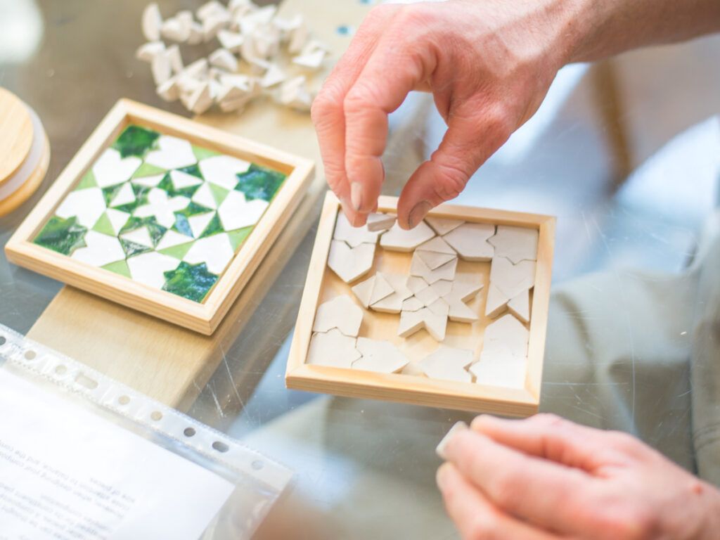 Hands fitting clay tiles into a coaster frame next to a finished green coaster