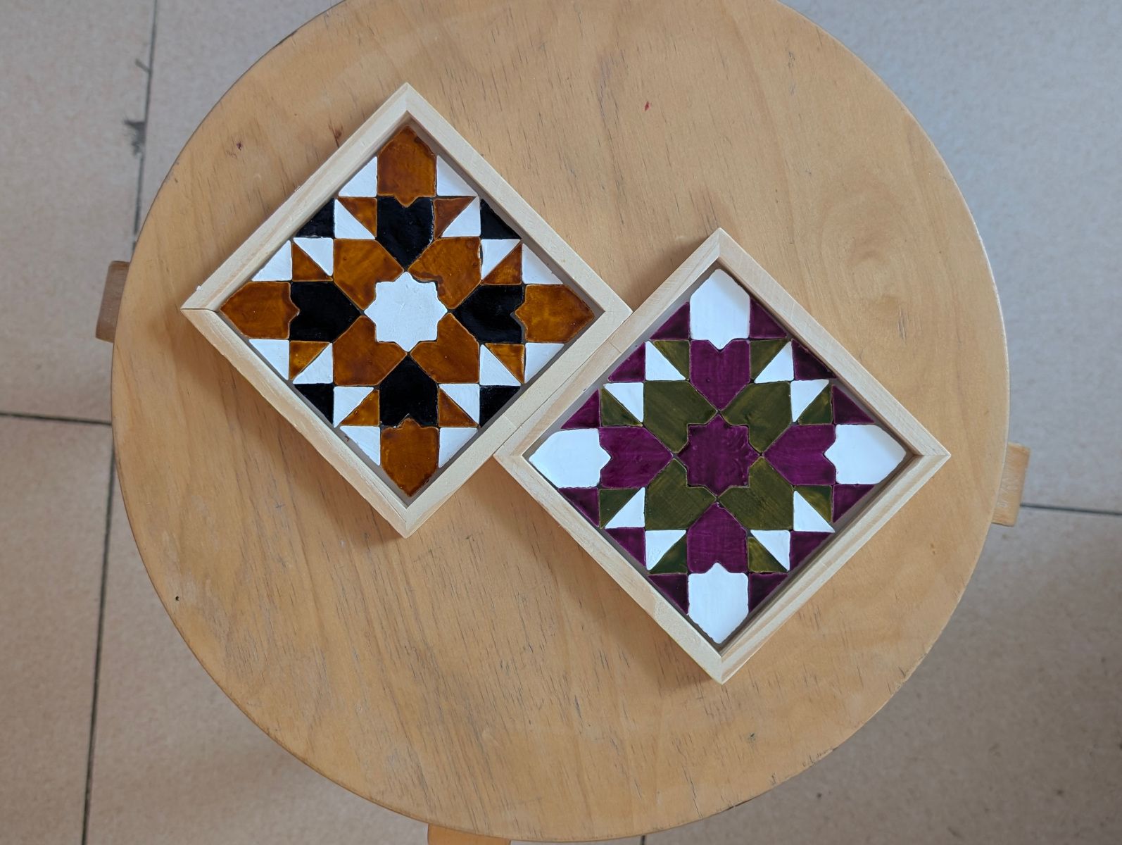 Two finished geometric mosaic coasters on a round wooden stool
